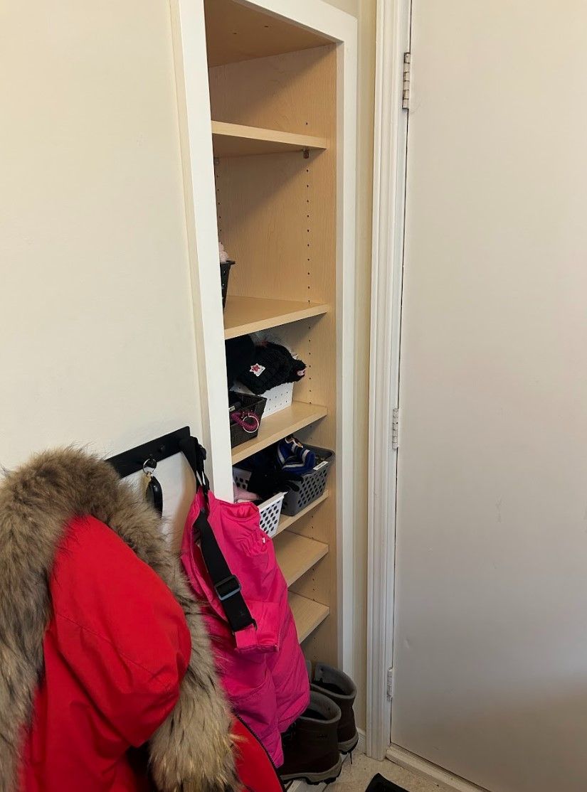 Narrow shelving unit built into a wall, holding various items like hats and baskets, next to a coat rack.