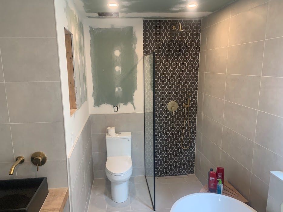 Bathroom renovation: grey tiled walls, mosaic shower, toilet, sink, and partially constructed features.