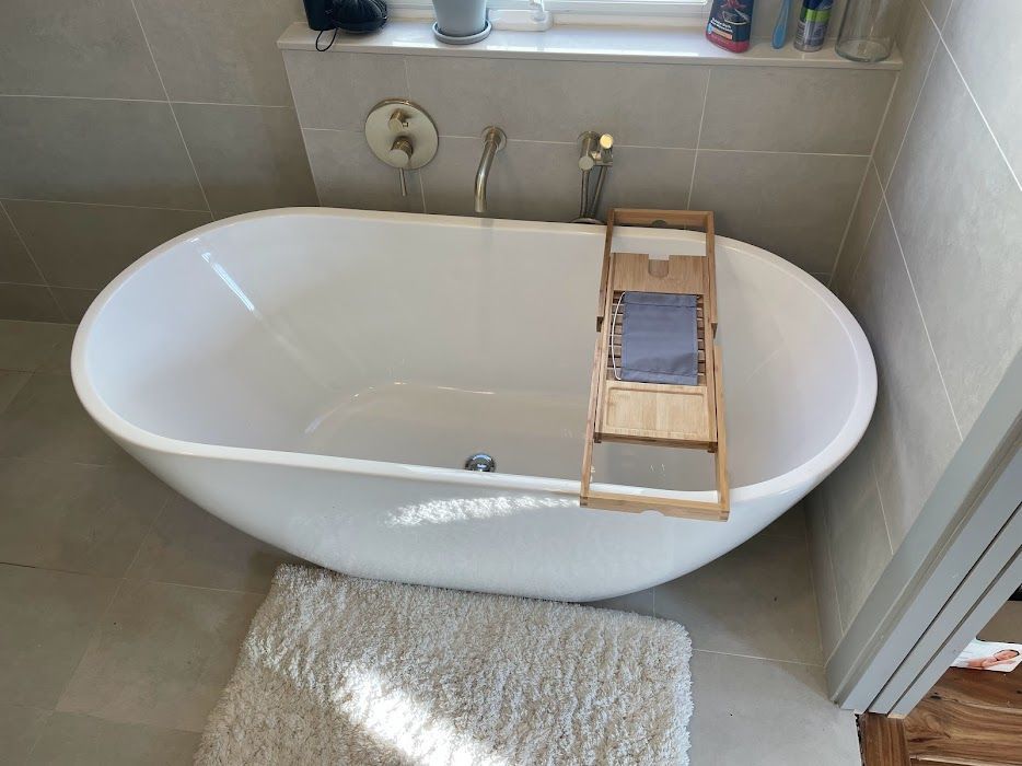 Oval white bathtub with wooden bath tray, in a light-tiled bathroom. A fluffy white rug lies on the floor.