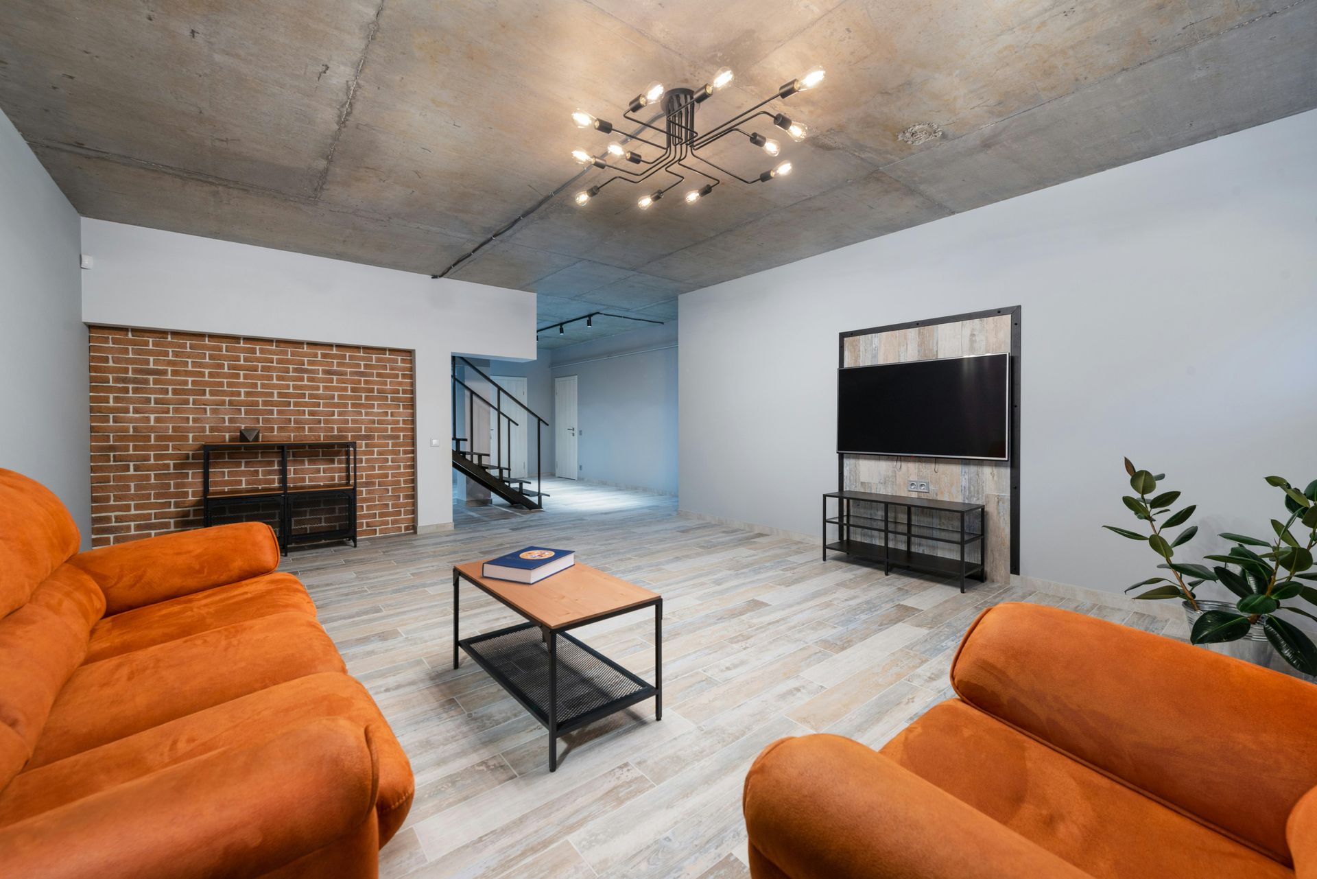 Modern living room with concrete ceiling, orange sofas, brick accent wall, and a TV on a dark metal stand.