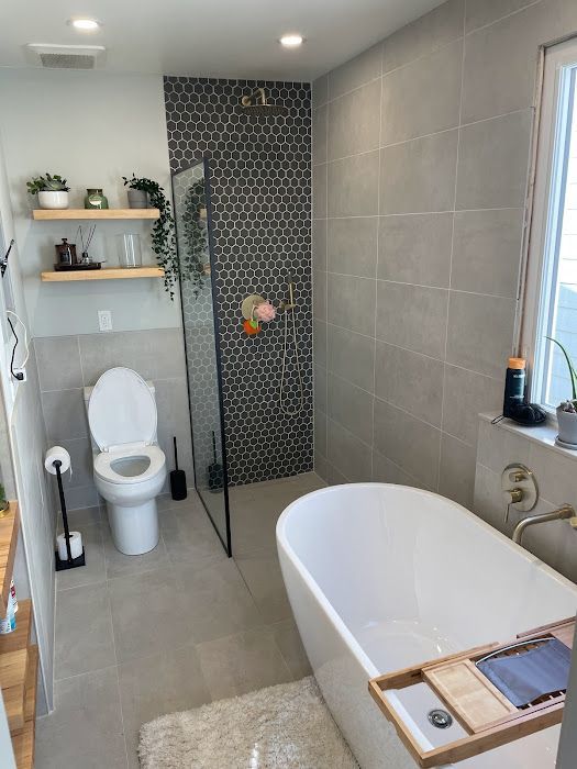 Modern bathroom with a white bathtub, toilet, and black shower, featuring gray and black tiles, and floating shelves.