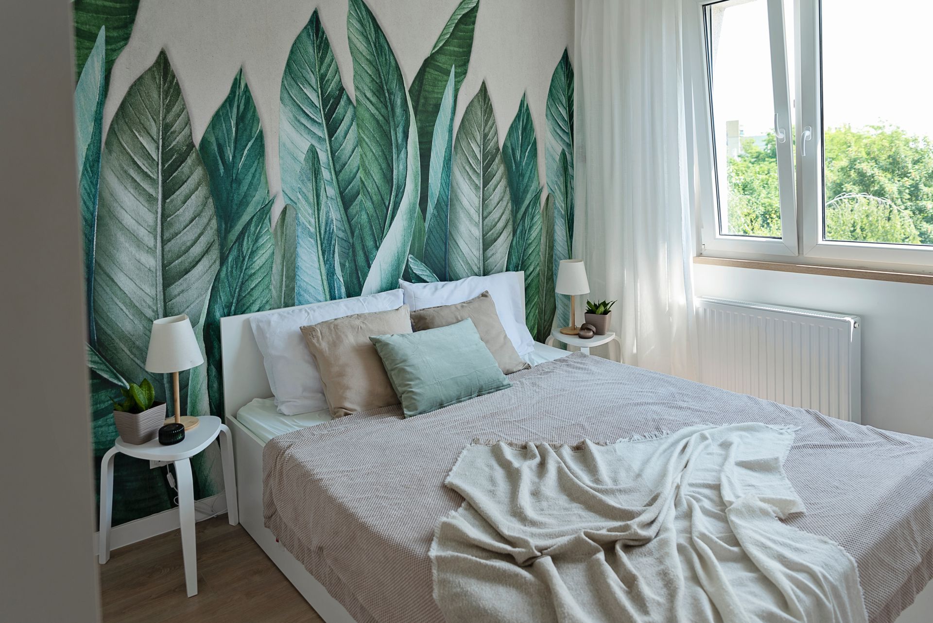 A bedroom with a bed , two nightstands , a window and a wall with leaves on it.