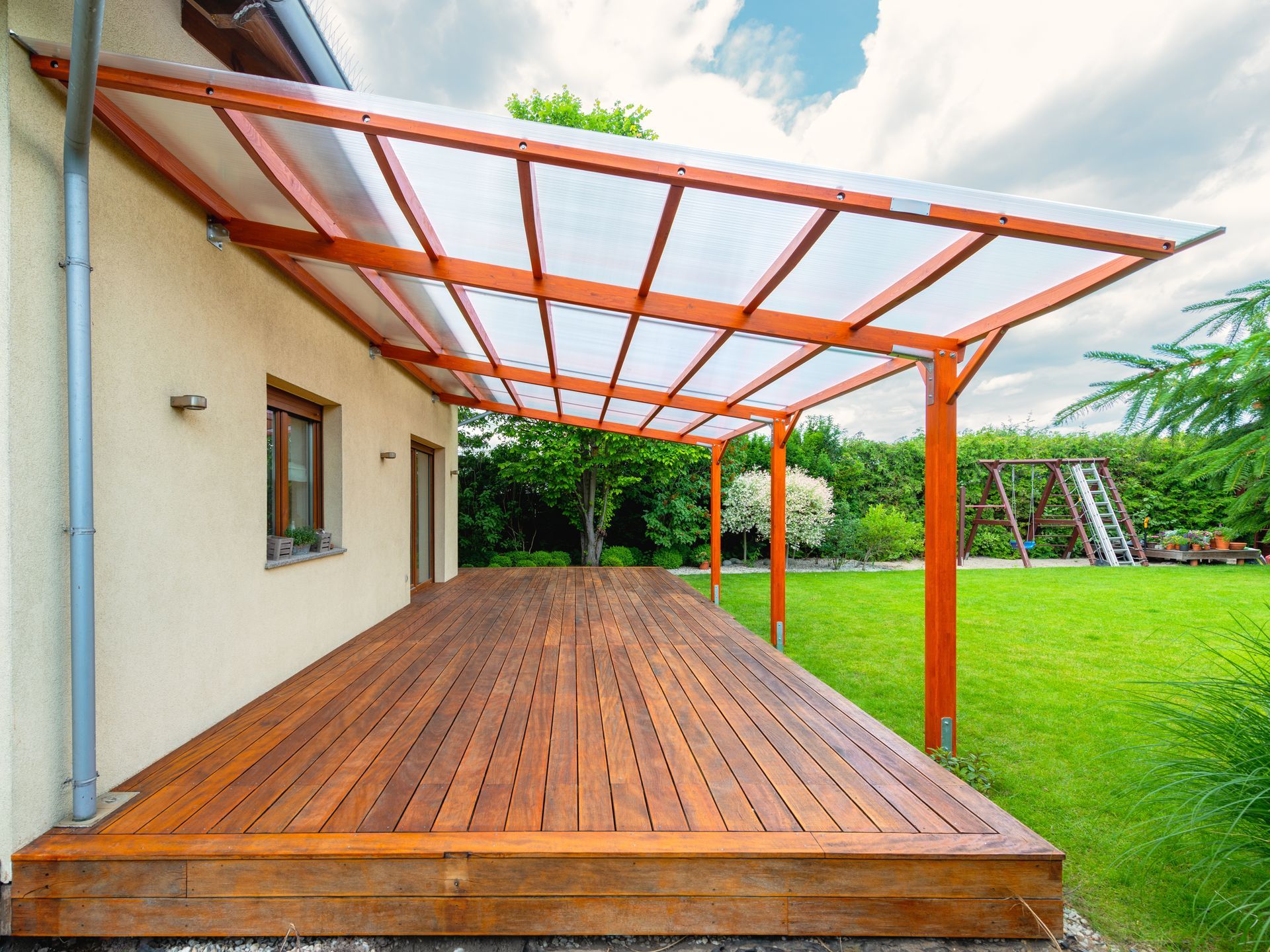 A wooden deck attached to a house with a translucent, sloped canopy overhead, overlooking a grassy backyard.