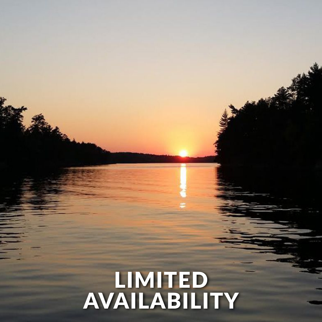 A warm, golden sunset glows over a calm lake, framed by silhouetted trees on both sides with a bright light reflection.