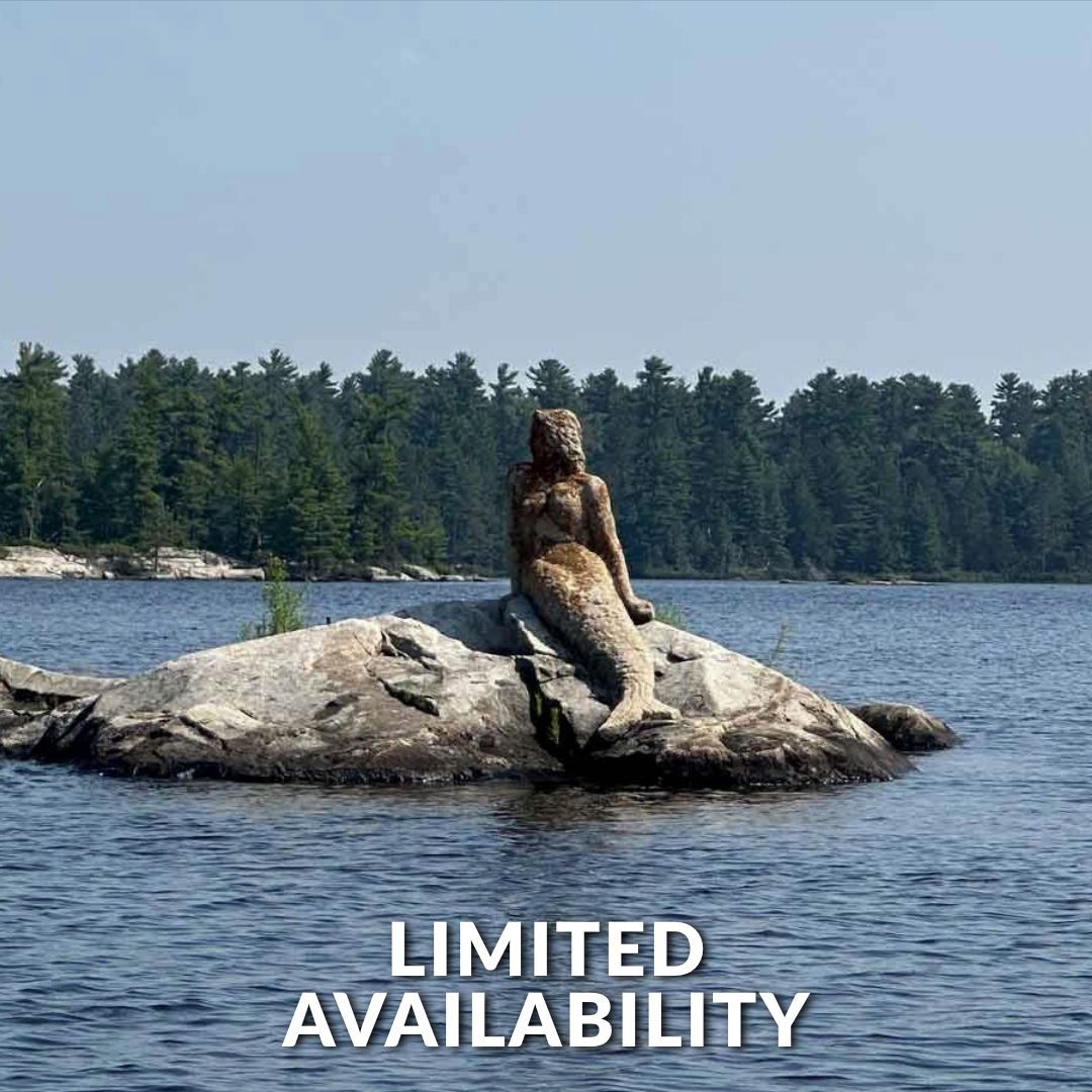 A mermaid sculpture sits on a rock in the middle of a calm, blue lake surrounded by a dense forest under a clear sky.