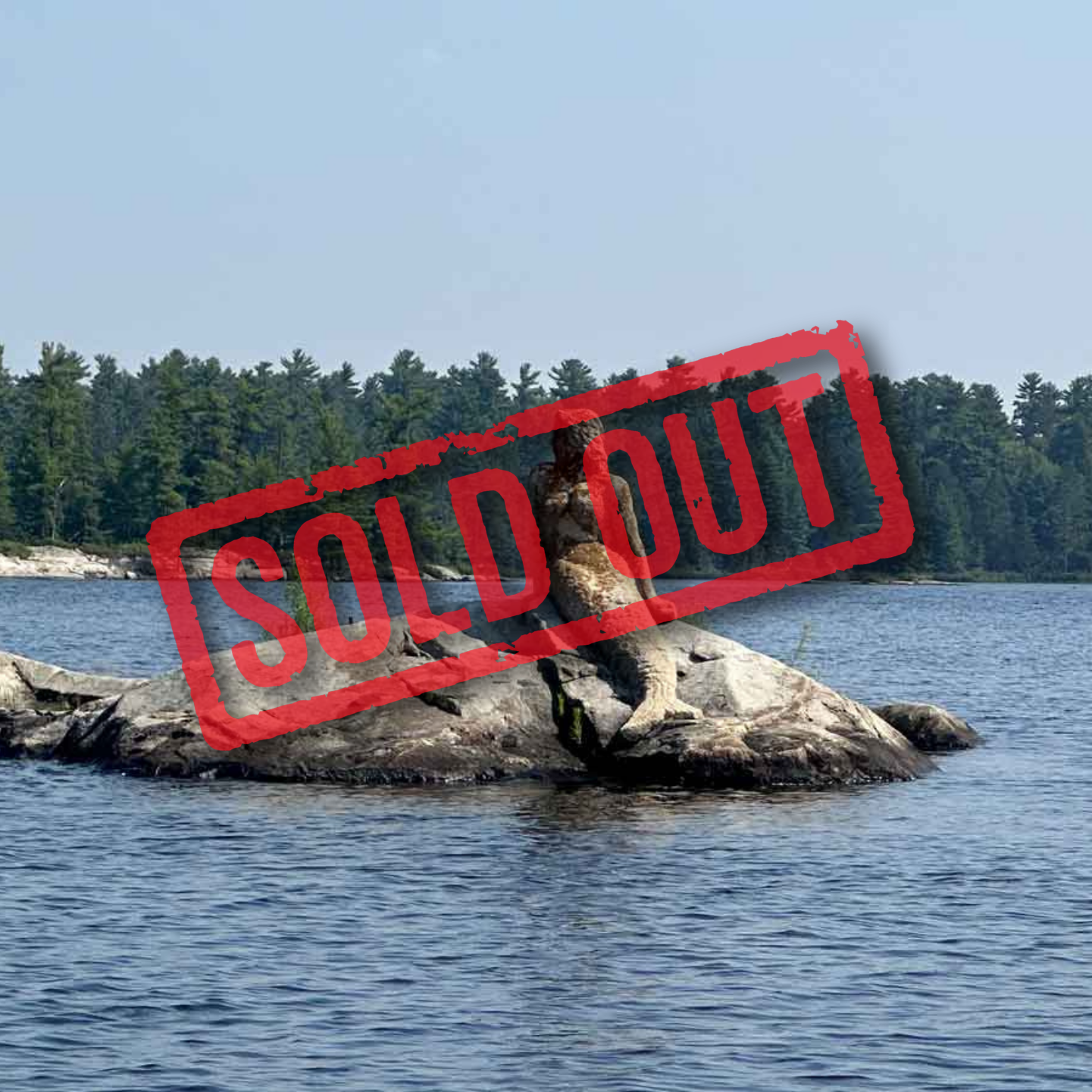 A mermaid sculpture sits on a rock in the middle of a calm, blue lake surrounded by a dense forest under a clear sky.