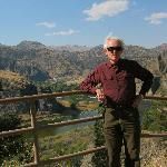 An older person in a maroon long-sleeved shirt and dark pants stands by a wooden railing overlooking a scenic valley.