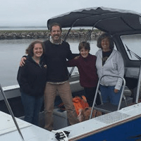 A family of four stands together smiling under the canopy of a boat on a cloudy day with a shoreline in the background.