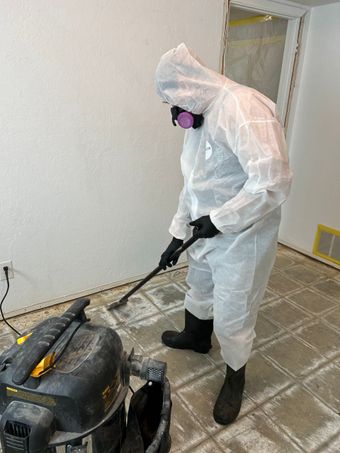 Person in protective suit vacuums debris from a tiled floor. Indoor setting, white walls, and respirator.