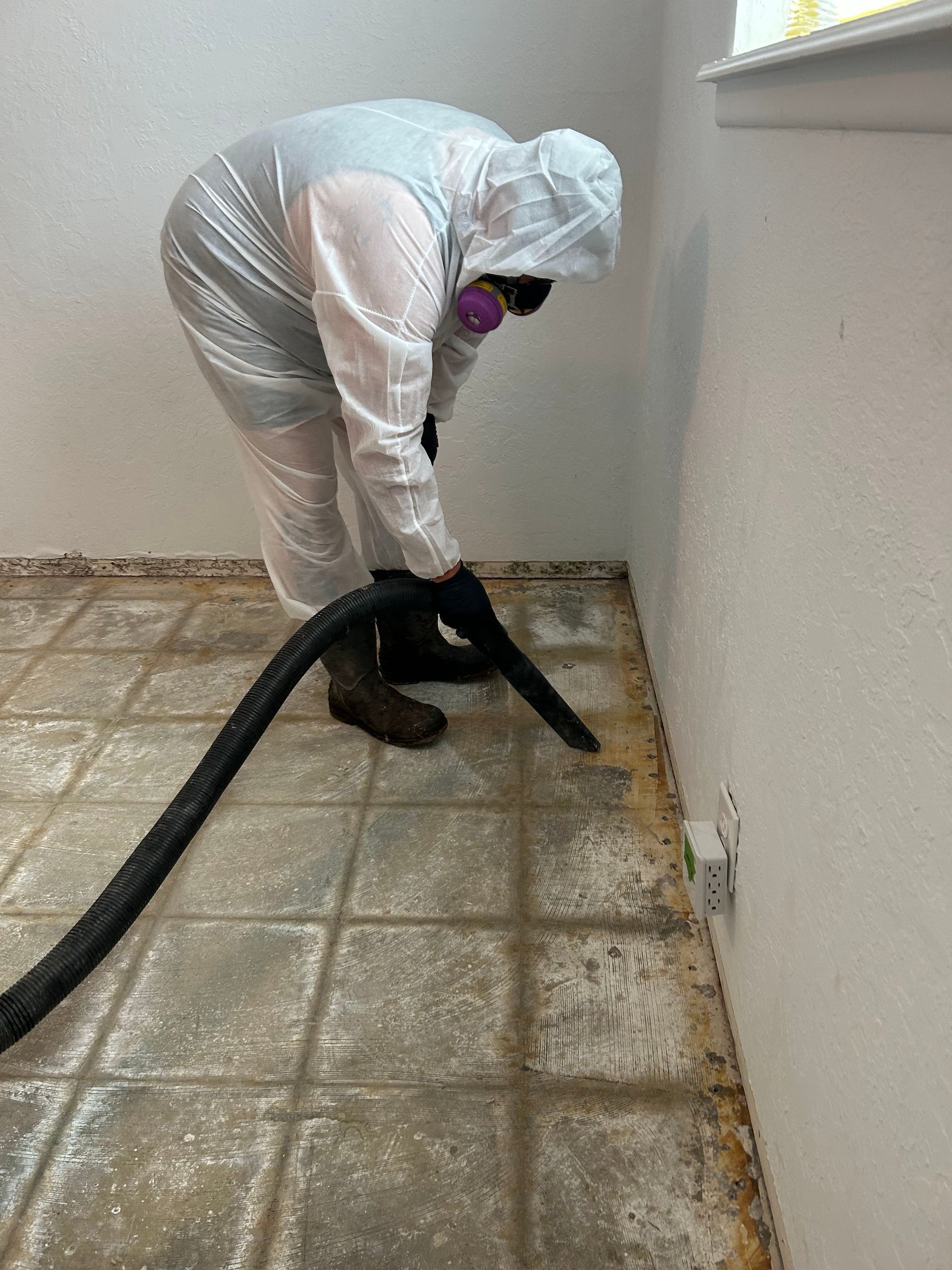 Person in hazmat suit vacuums a dirty tiled floor in a room; setting may be a house, with a white wall.