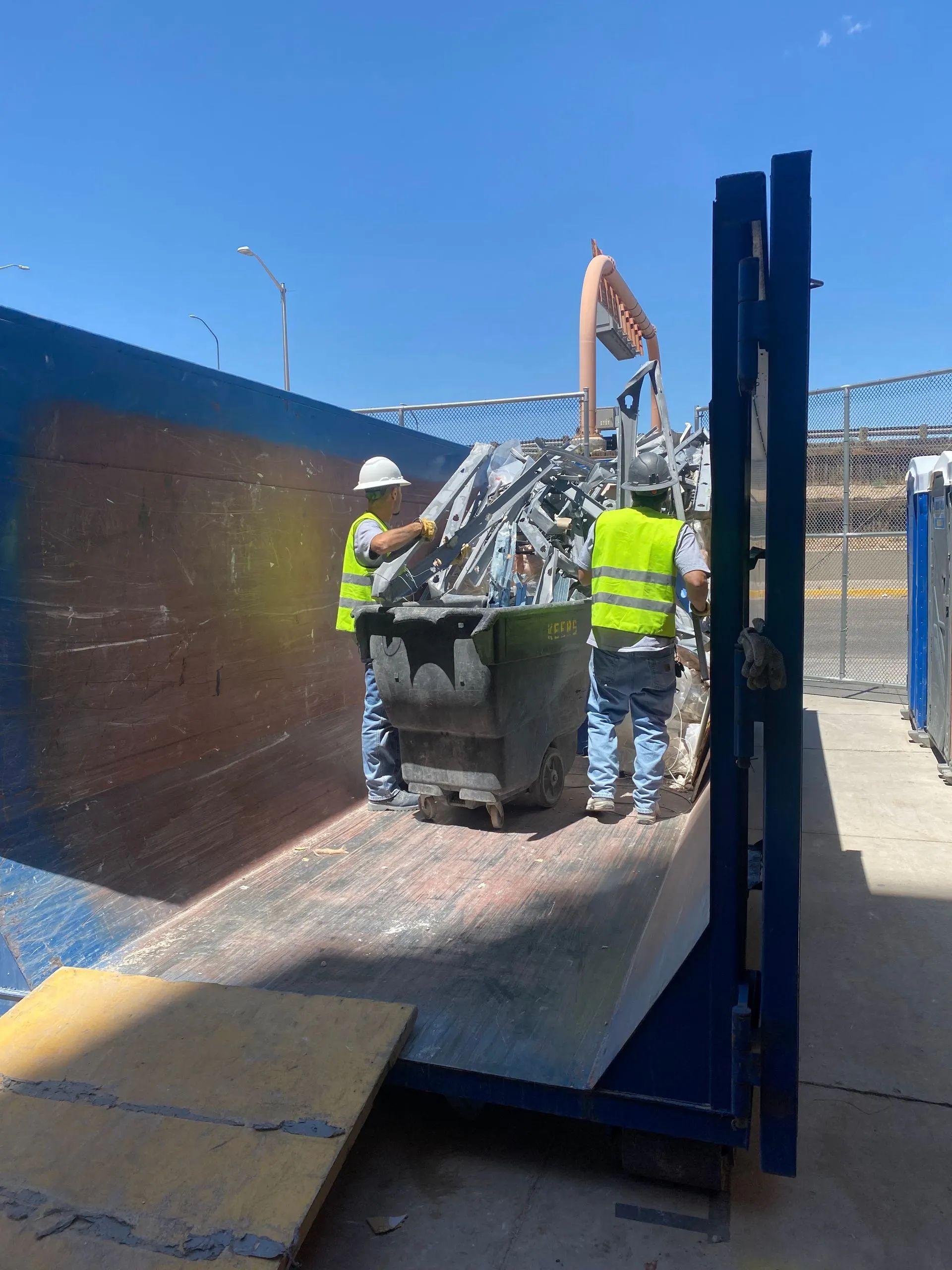 Two workers loading metal scraps into a dumpster on a construction site.