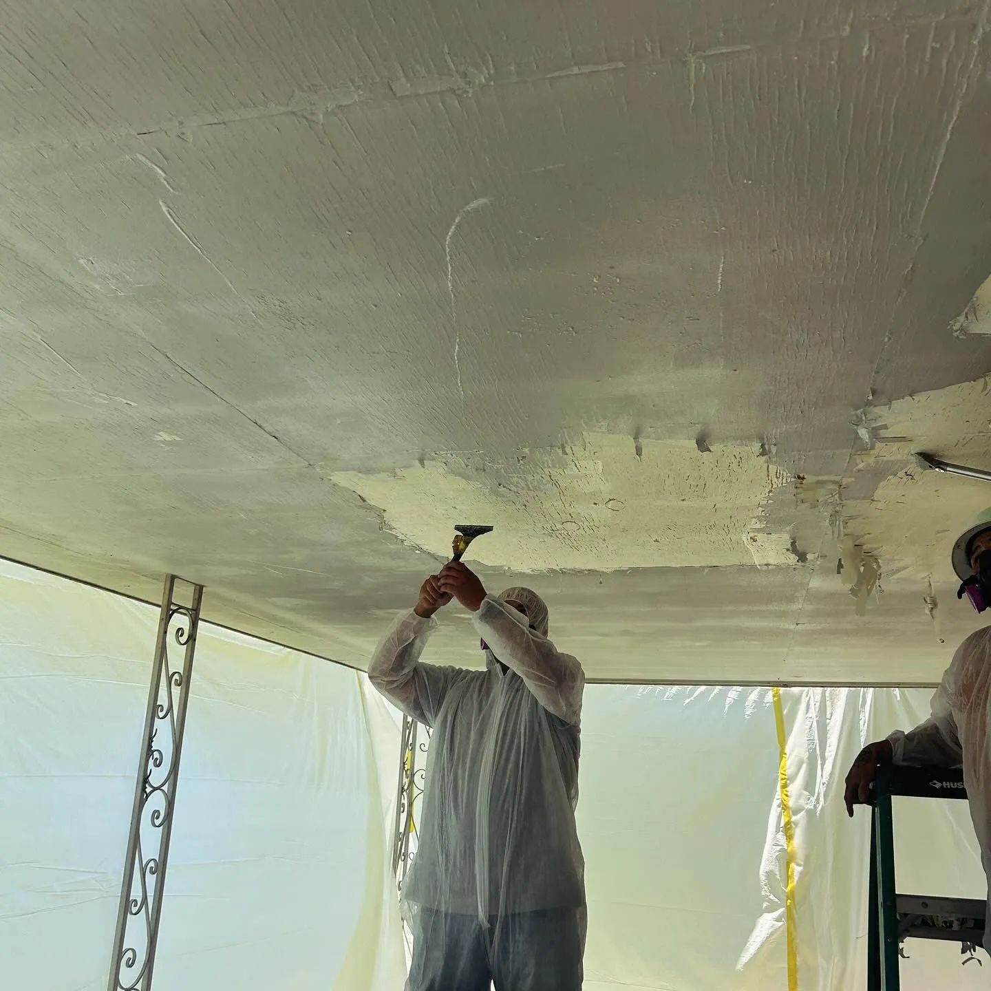 Person in protective suit applying coating to a ceiling with a scraper, outdoors.