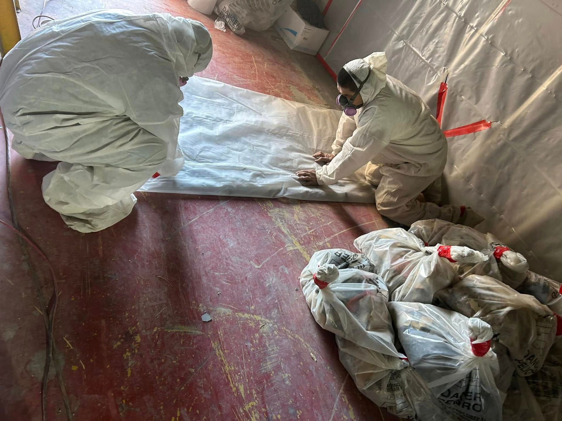 Two people in protective suits removing material from a red surface, bags of debris nearby.