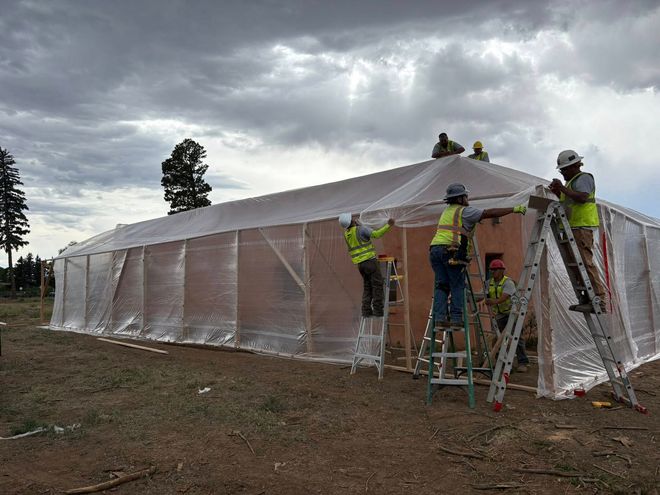 Construction crew installing plastic sheeting on a long, low building under a cloudy sky.