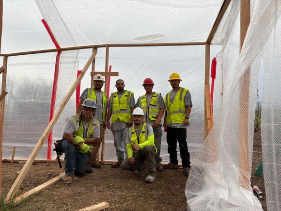 Construction workers pose inside a wooden frame covered with plastic sheeting, wearing vests and hard hats.