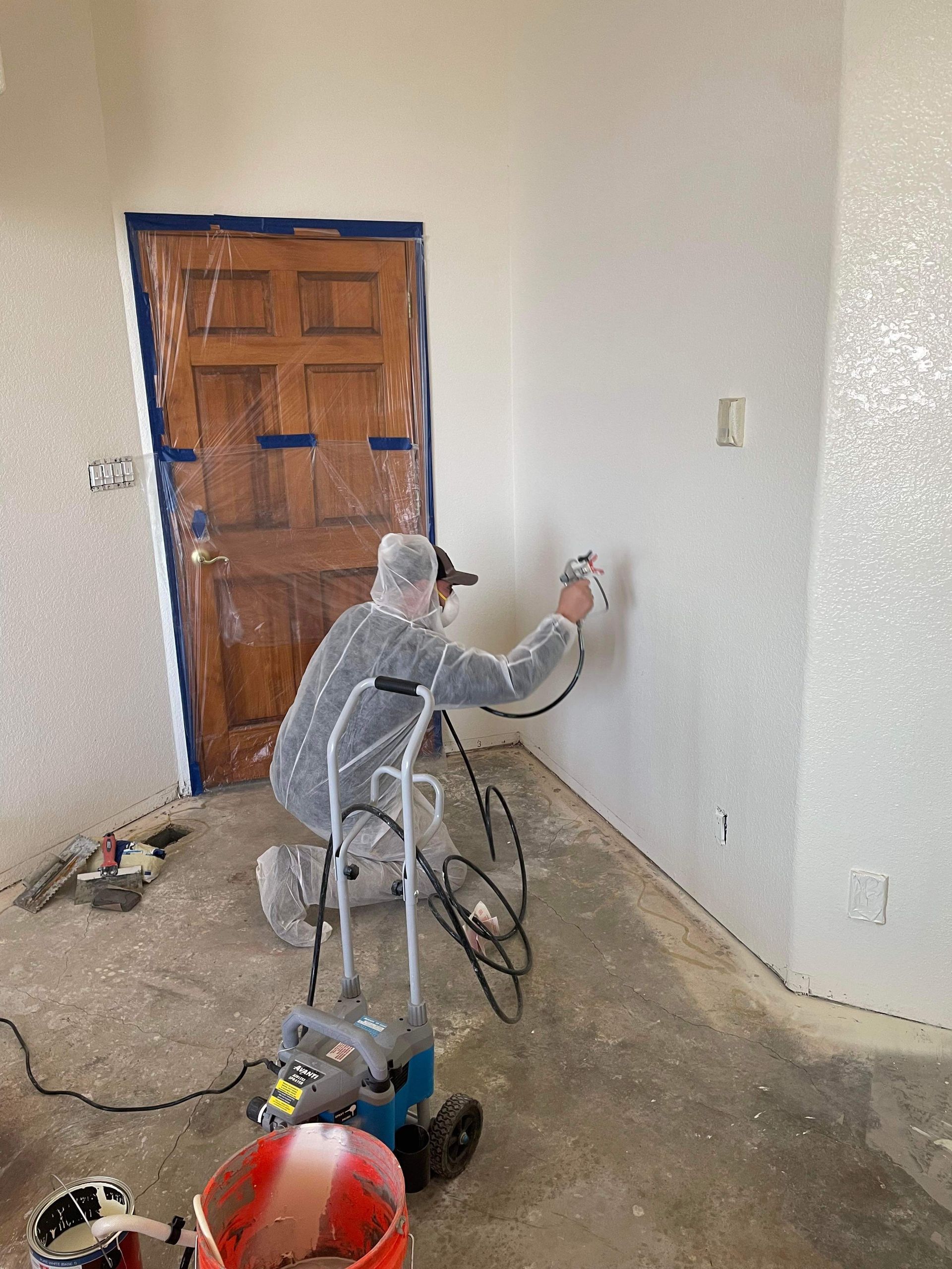 Person spraying texture on a wall indoors. Door and floor are covered, sprayer and bucket on the floor.