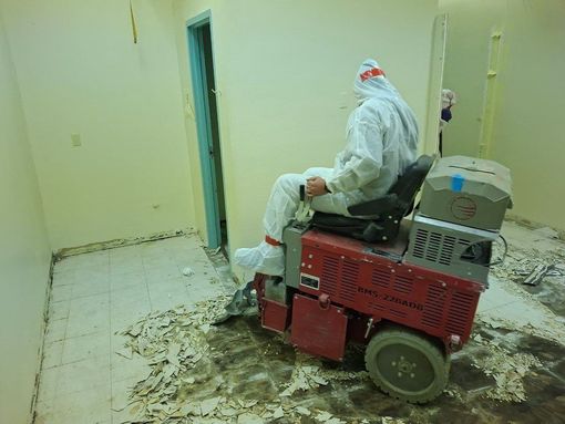 Person in protective suit operating a floor scraper, removing tile in a room.