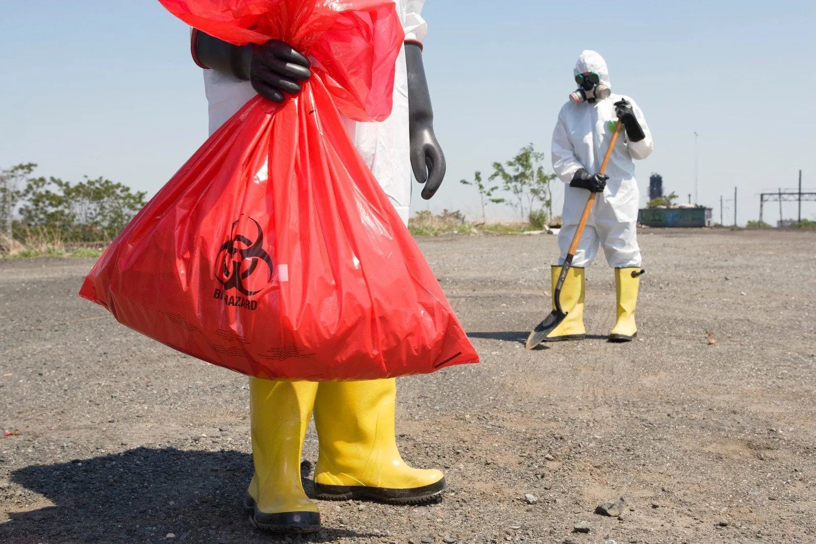 Workers in protective suits carrying a red biohazard waste bag at an outdoor cleanup site