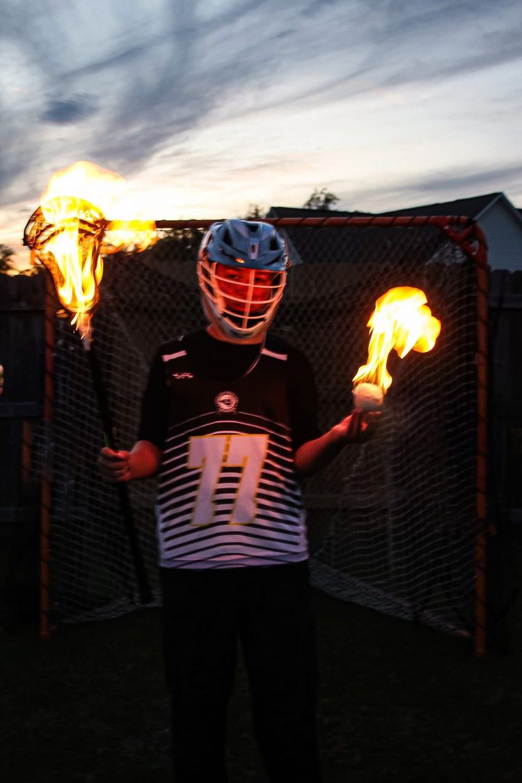 Lacrosse player with flaming stick and ball, standing in front of goal at dusk.