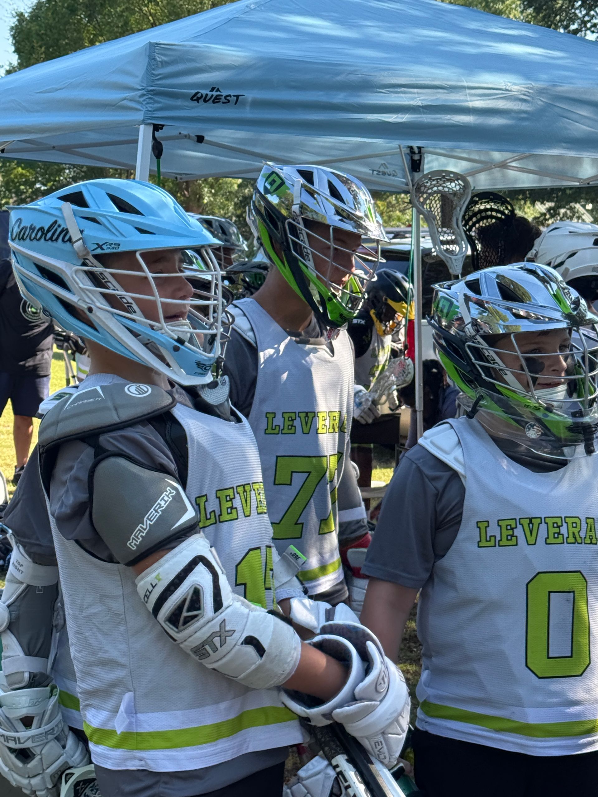 Lacrosse players wearing helmets and jerseys, standing under a canopy.