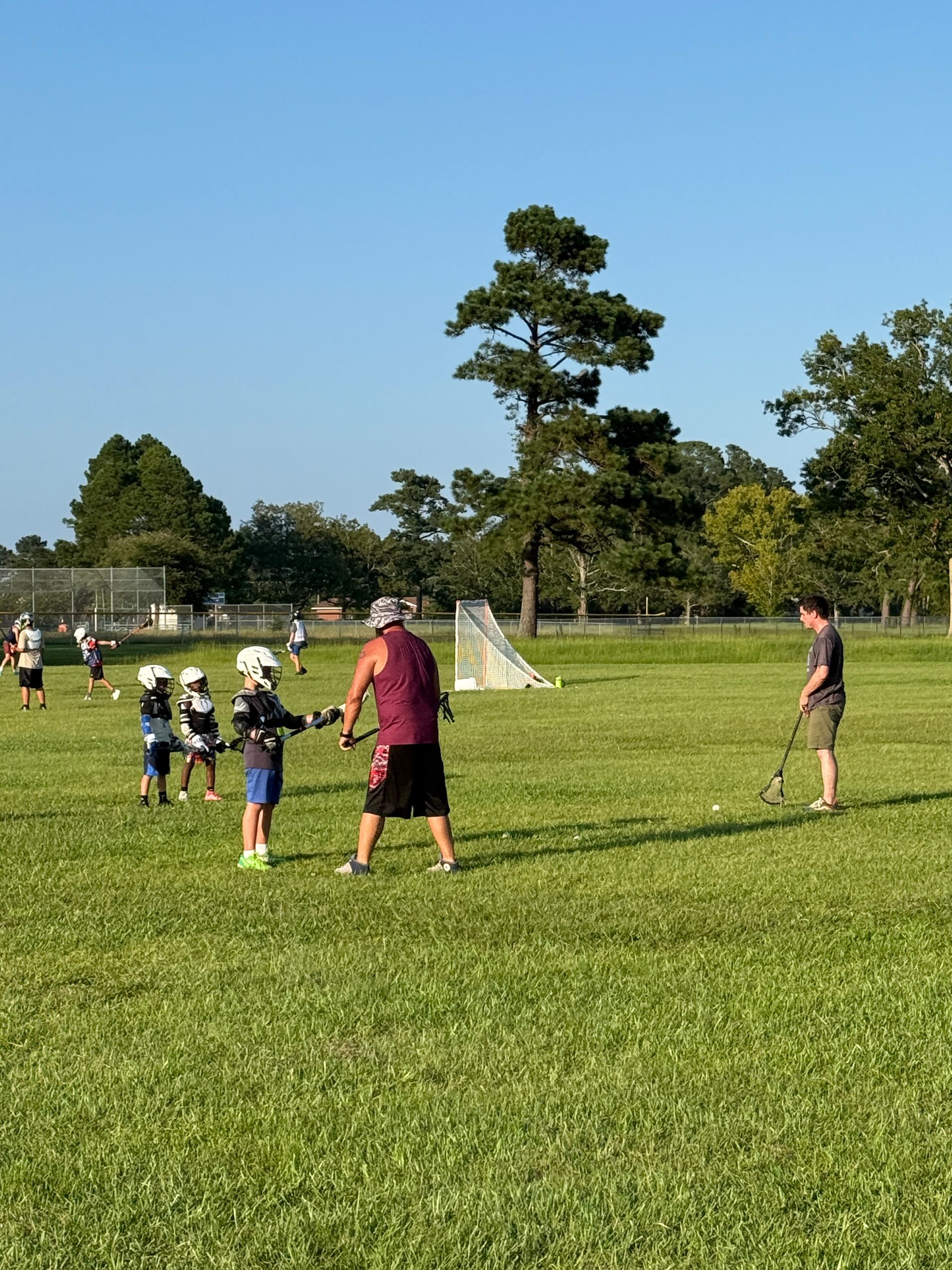 Lacrosse practice on grassy field. Kids in helmets and coach demonstrating stick handling. Goal in background.