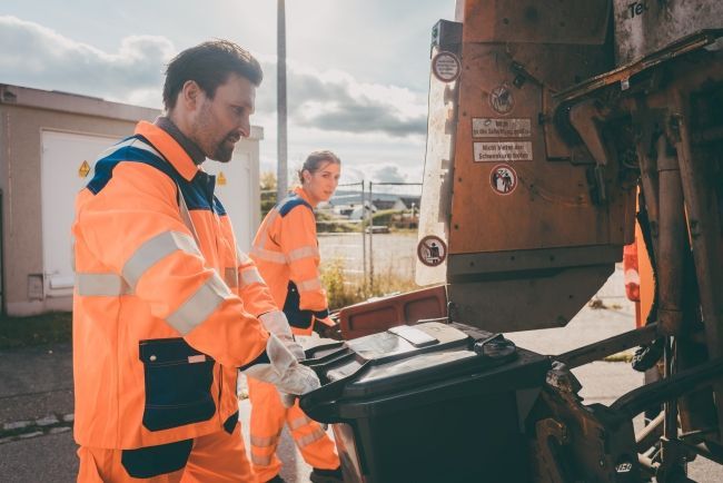 Two workers in safety gear loading a trash bin into a collection truck.