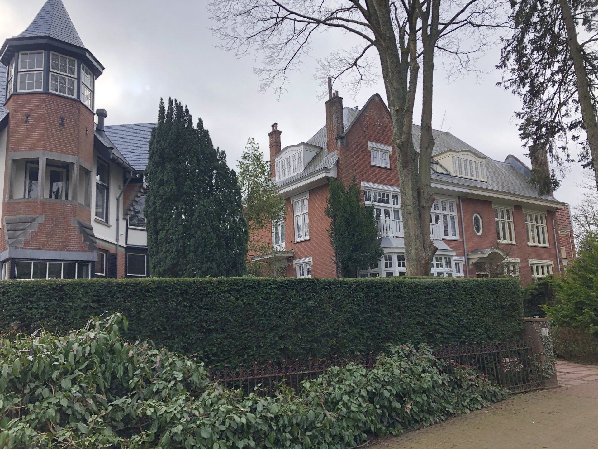 Een groot bakstenen huis met een toren en een heg ervoor.