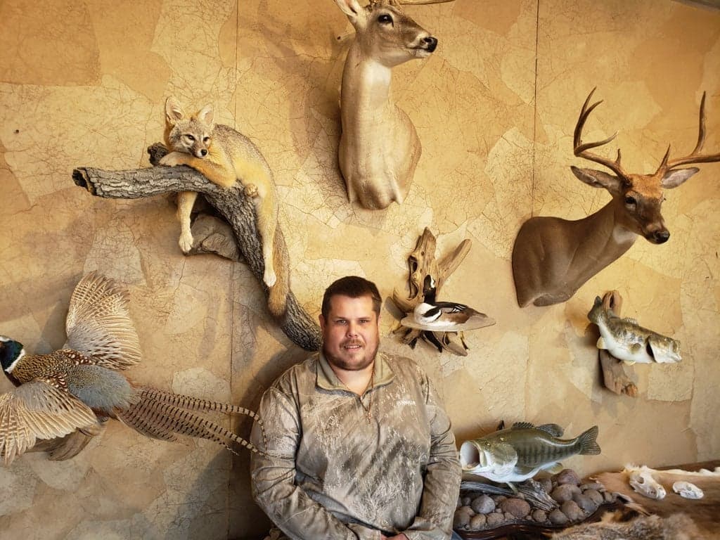 Man in camo posing next to taxidermied animals on a textured wall. Deer, fox, fish, and birds.