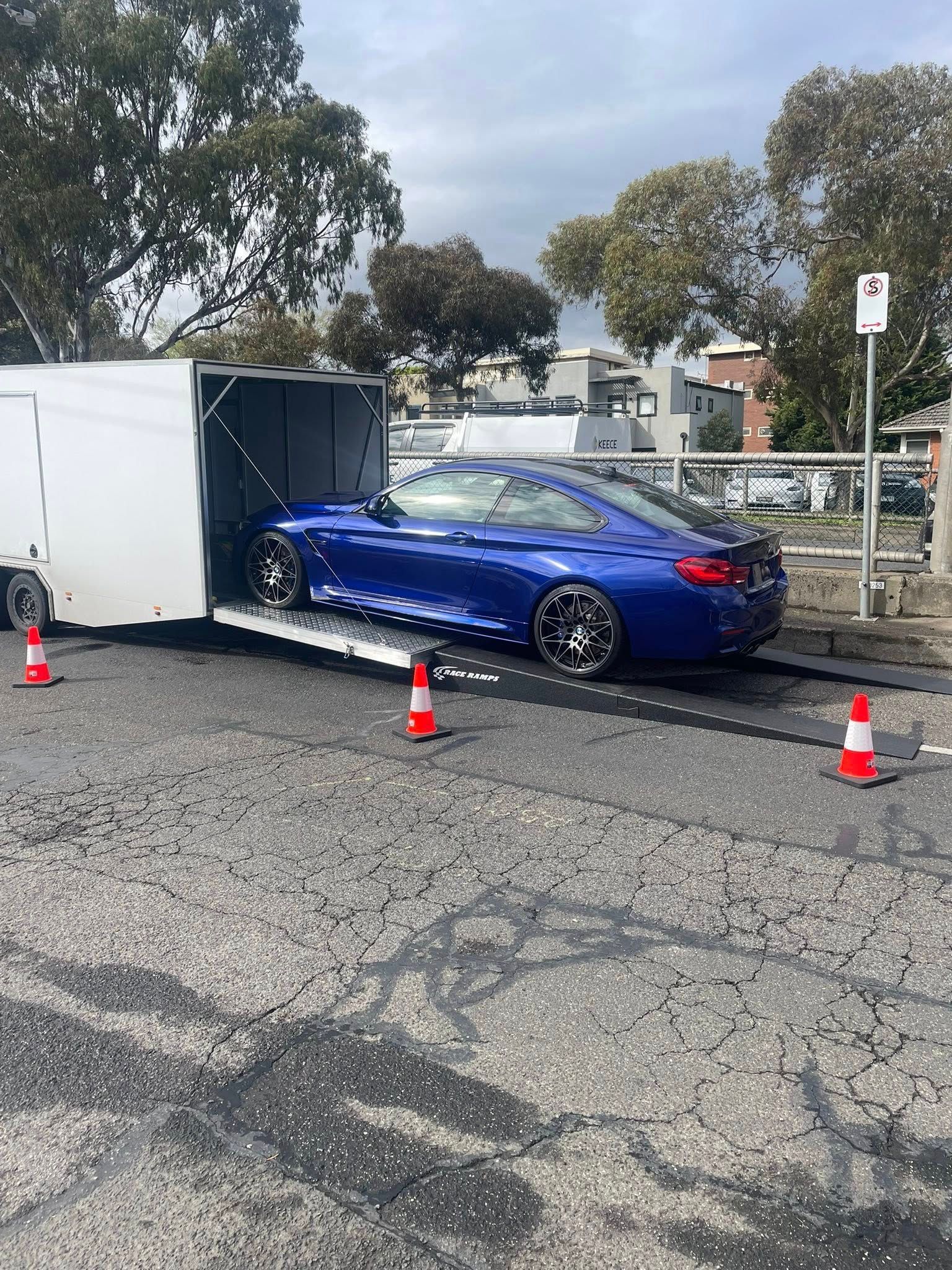 Blue car exiting a white trailer on a cracked asphalt road, with traffic cones and trees in the background — Full Throttle Enclosed Car Transport in Kilmore, VIC