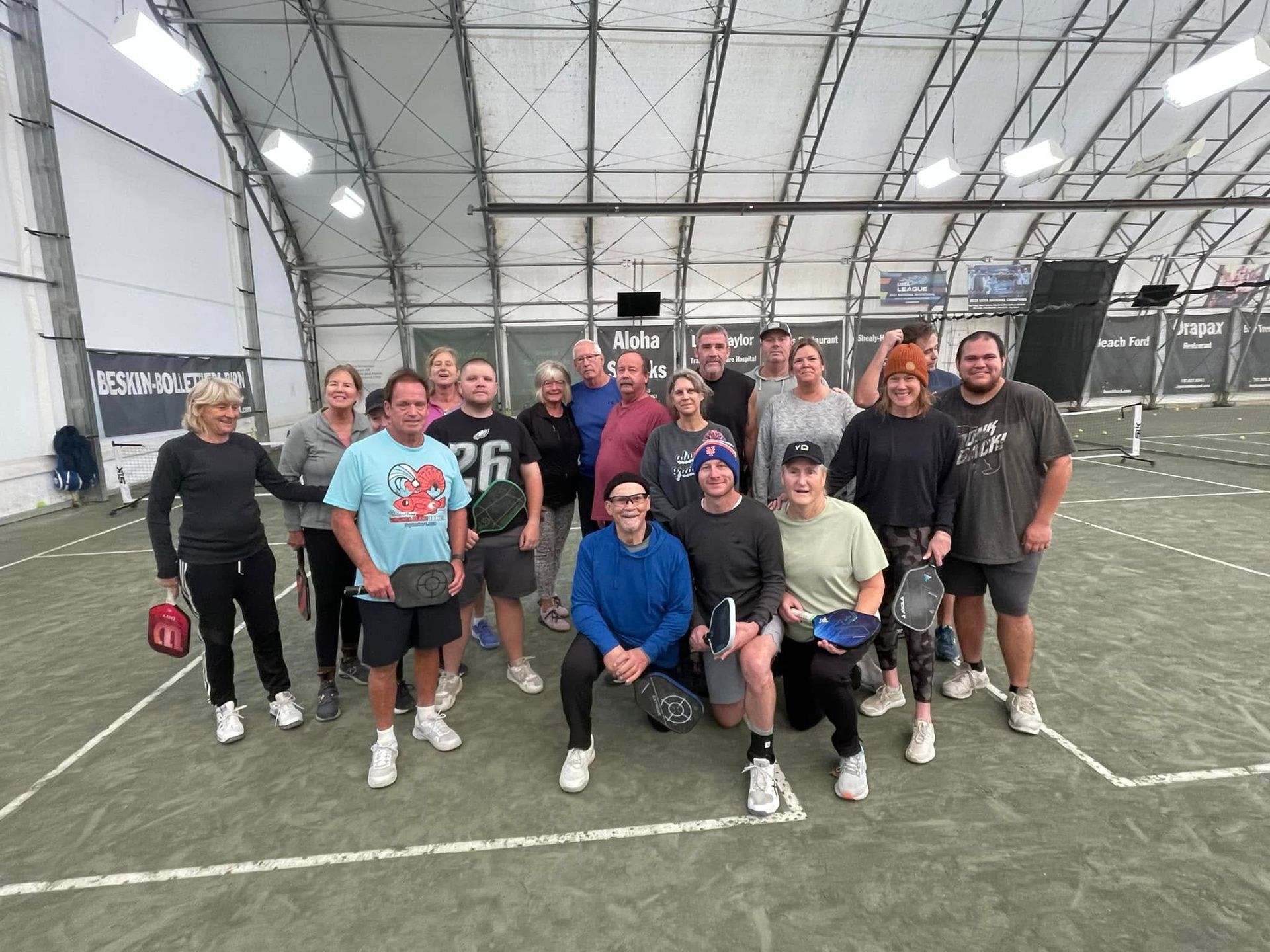 Group of people posing on a pickleball court indoors. Some hold paddles, smiling.