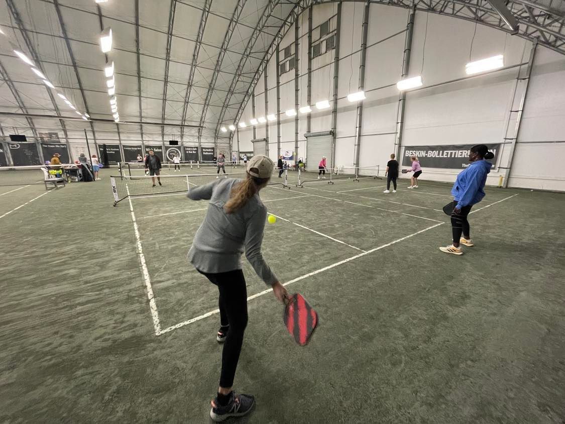People playing pickleball indoors on a green court; one woman hits the ball.
