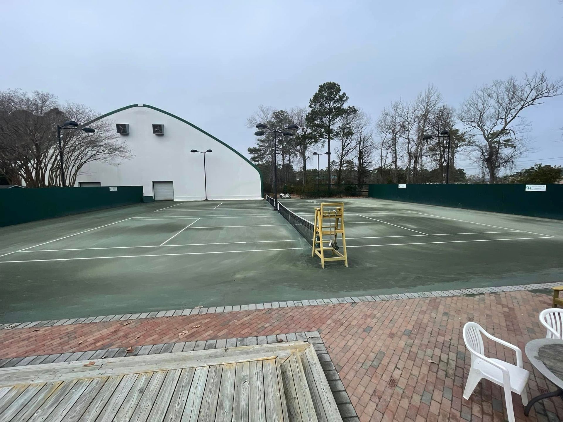 Tennis court with green surface, white lines, and net. Building and trees in the background, chairs in the foreground.