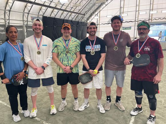 Six people with medals, pose on a court. Some hold paddles, smiling. Indoors, under a white dome.