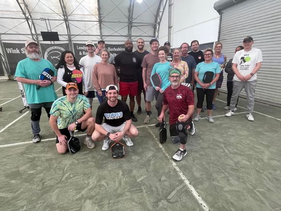 Group of people at a pickleball court, posing. Some hold paddles, and wear athletic clothing, indoors.