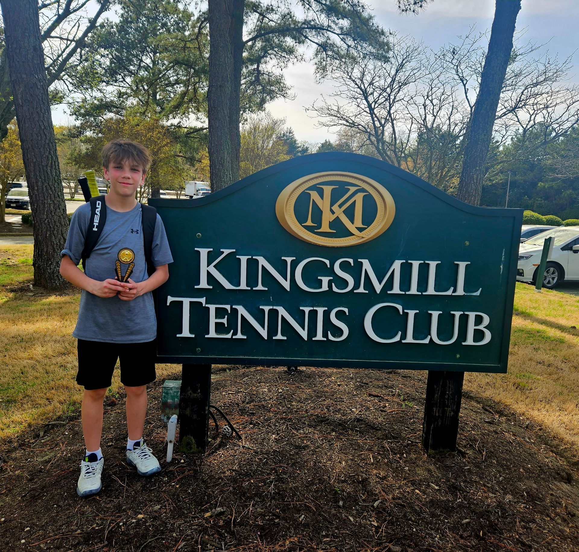 Boy with trophy stands in front of the Kingsmill Tennis Club sign, smiling.
