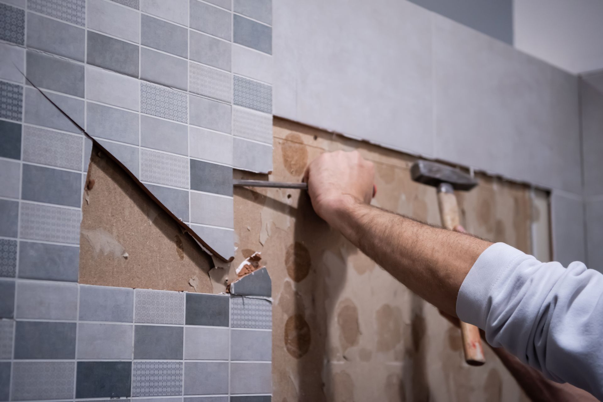 A man is removing tiles from a wall with a spatula and hammer.