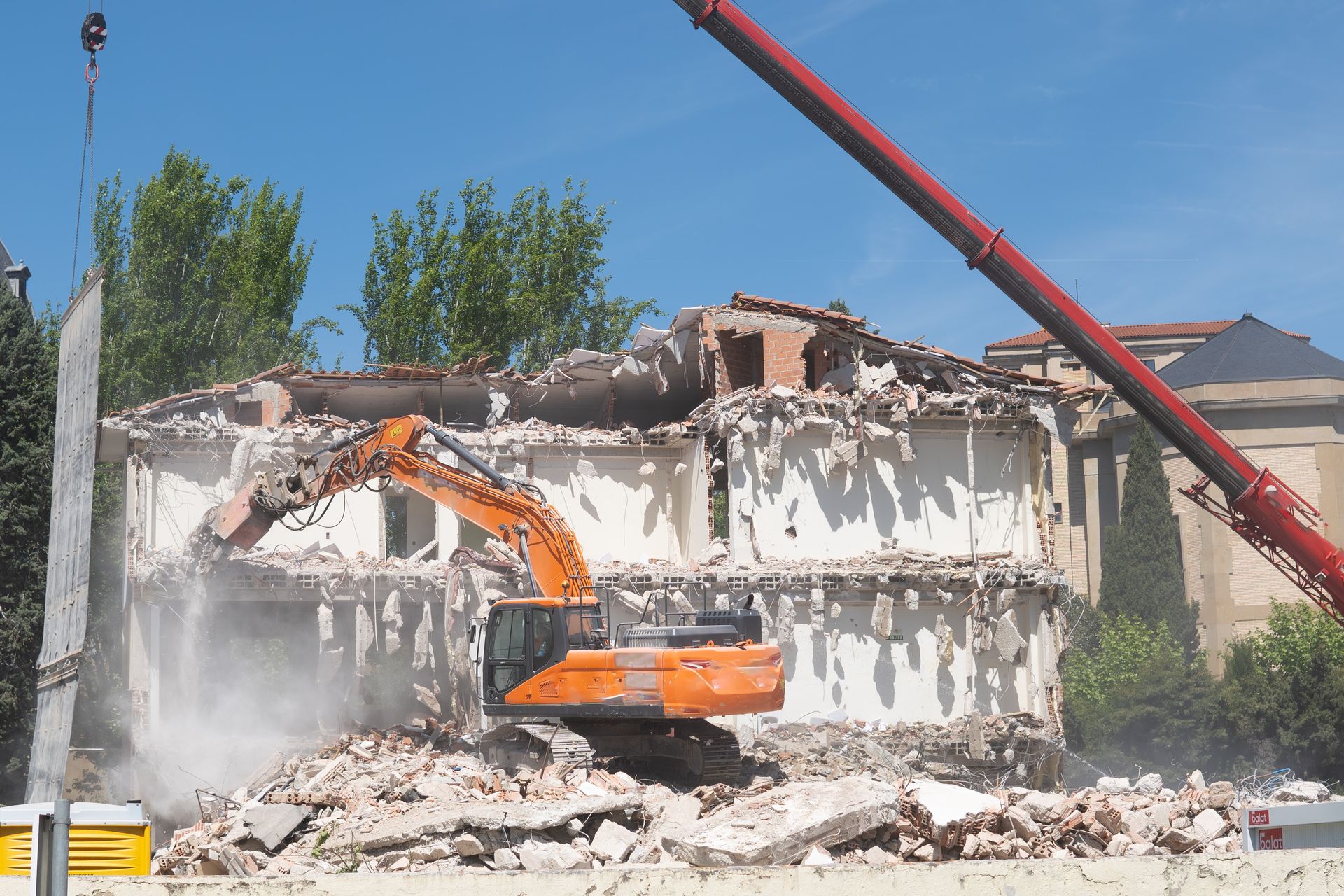 An excavator is demolishing a building with a crane in the background.