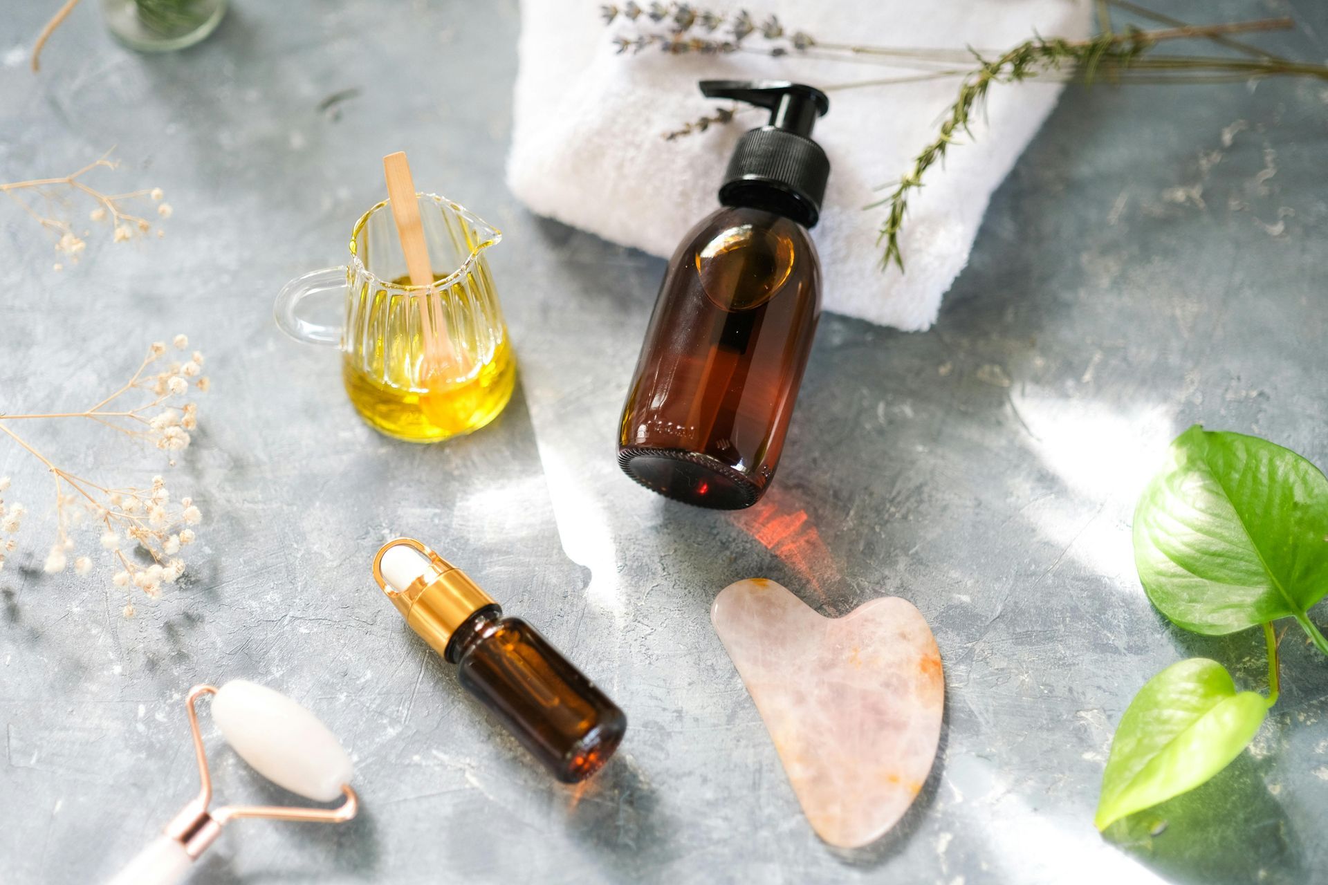 Skincare products: bottle, oil, roller, and Gua Sha tool on a grey surface with a towel and plant.