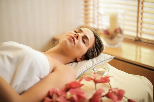 Woman relaxing at a spa, eyes closed, draped in white, with flowers, candle, and light coming through blinds.