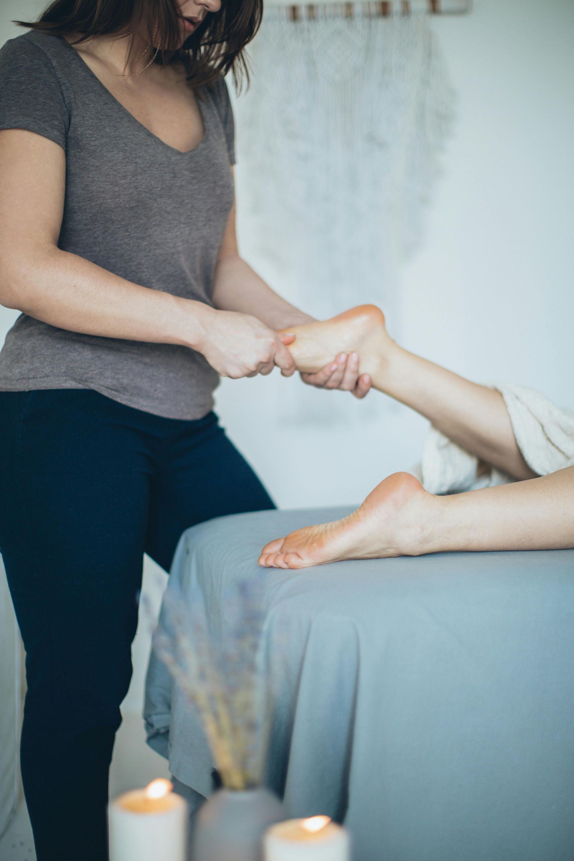 Woman giving foot massage on a massage table; candles and vase with dried flowers in front.