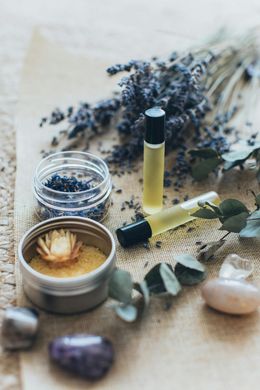 Lavender-themed self-care items: roller bottles, bath salts, dried flowers, crystals on a burlap cloth.