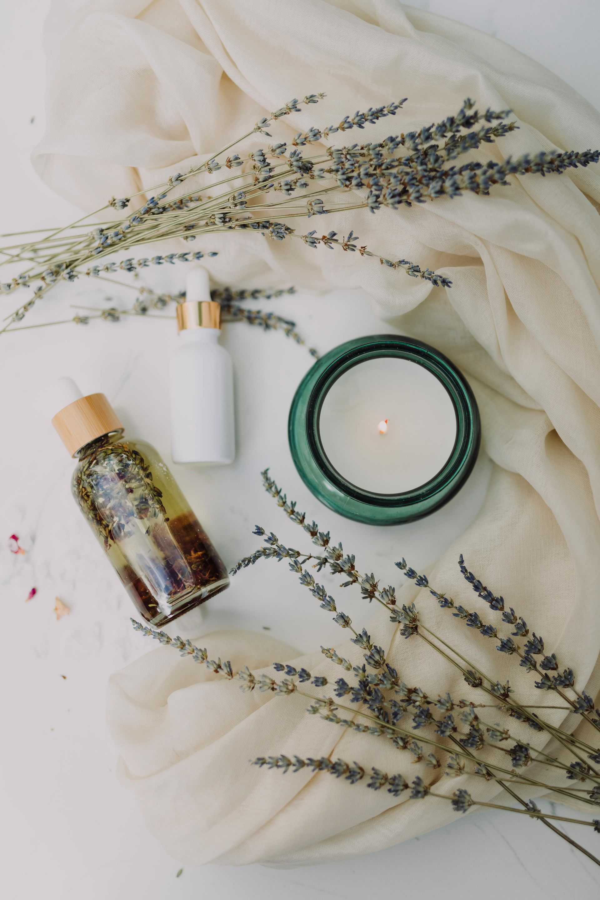 White bottles, green candle, lavender sprigs, and beige fabric on white background.