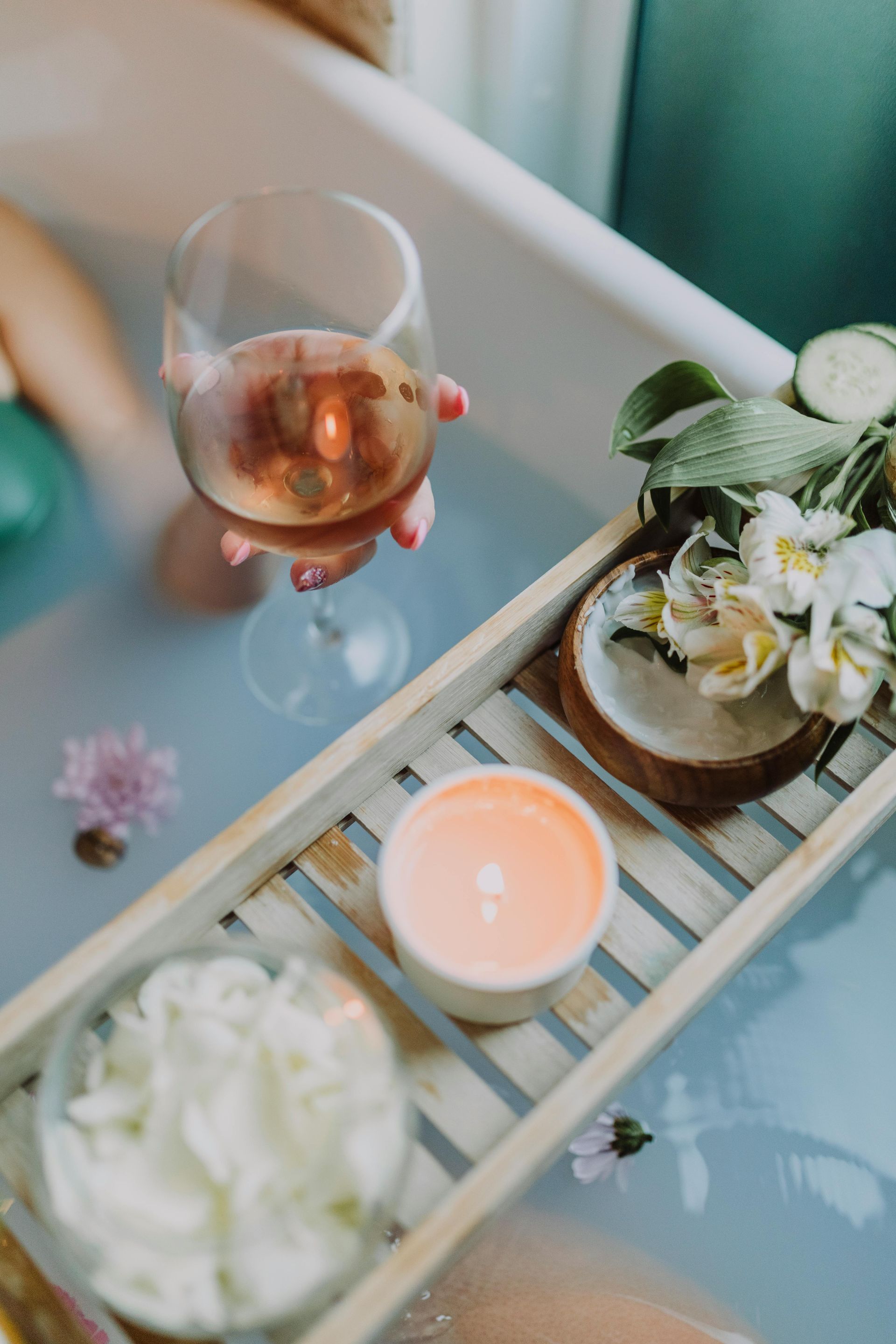 Person relaxing in a bathtub, holding wine glass. A wooden tray holds a candle, flowers, and coconut.