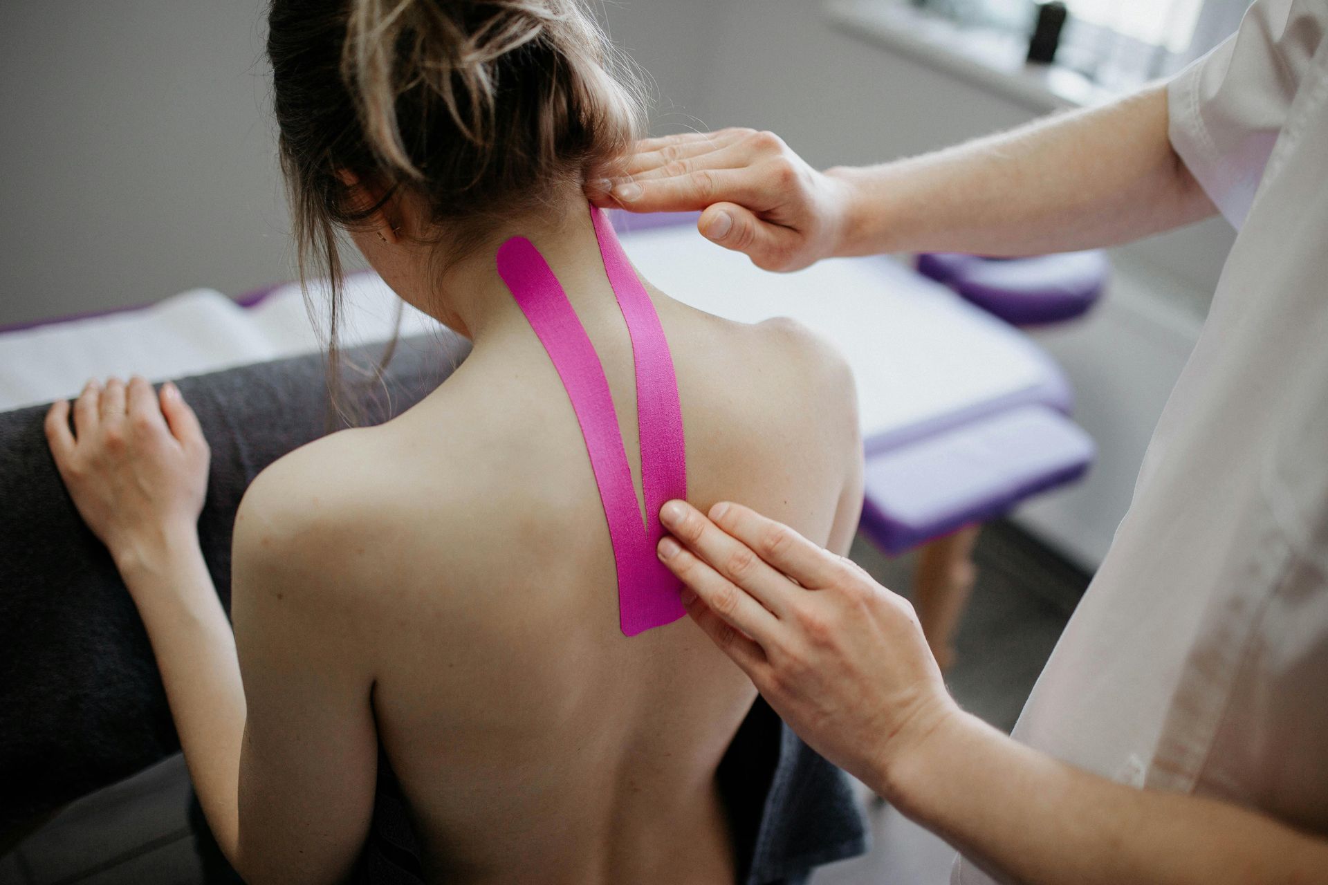 Hands applying pink kinesiology tape to a woman's upper back and neck in a treatment room.