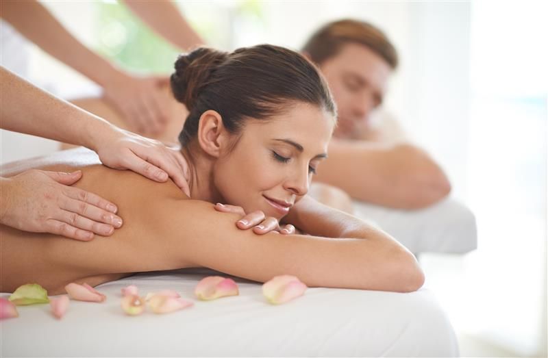 Woman receiving back massage at a spa with another person in the background, white sheets, rose petals.