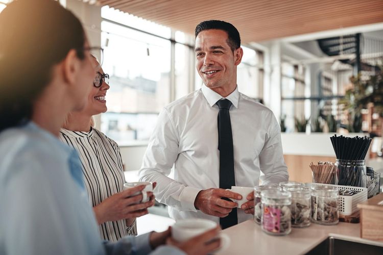 Three people at a coffee bar, smiling and holding mugs. Man in a white shirt, two women, bright interior.