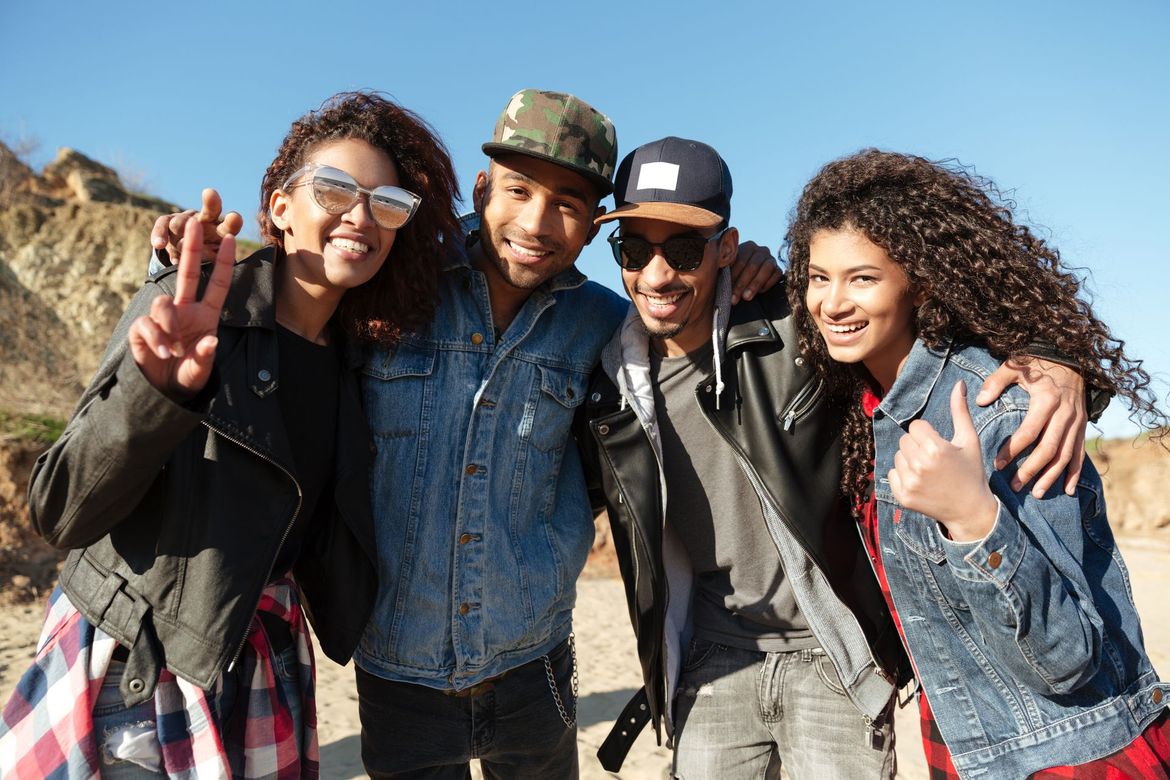 Four young friends in denim and jackets, smiling and making gestures, outdoors.