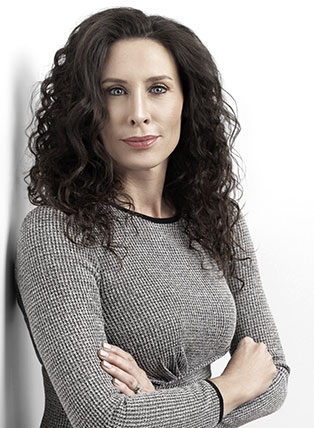 Woman with dark curly hair, arms crossed, wearing a gray dress, leaning against a white wall.