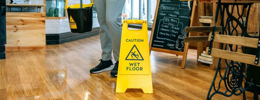 A person with a yellow wet floor sign in a wooden floor cafe, and mop bucket.