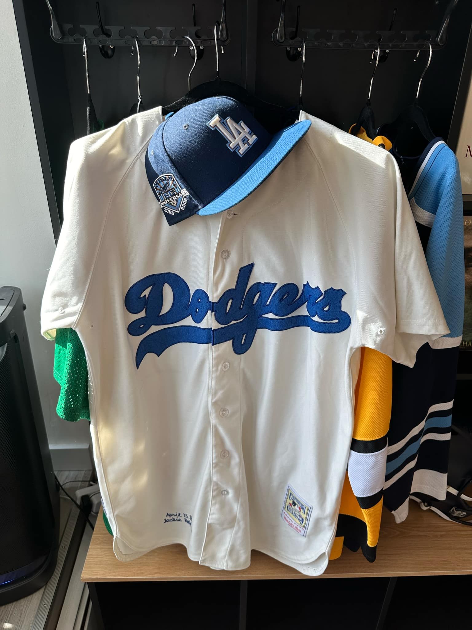 A white dodgers baseball jersey is hanging on a hanger next to a hat.